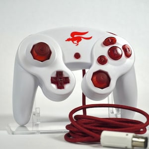 May include: A white video game controller featuring red translucent buttons, a red directional pad, and a red analog stick. A red graphic of a running fox is printed on the center. The controller includes a long, braided red cable with a white connector.