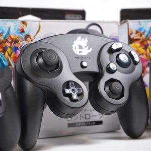 May include: A black wired video game controller featuring a white Super Smash Bros. flame logo on the center. The controller includes a directional pad, two analog sticks, and multiple action buttons. It is positioned in front of its original retail packaging.