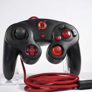 May include: A black wired video game controller featuring a red braided cable and a red lightning bolt logo at the top center. The device includes a black analog stick, a red directional pad, red action buttons, and a red C-stick.