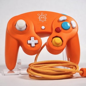 May include: An orange wired video game controller featuring a white joystick, a white directional pad, a large blue A button, a red B button, and white X and Y buttons. A white sun-shaped logo is printed above the center start button.