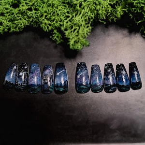 Peut inclure: Un ensemble de dix faux ongles avec un motif de ciel nocturne peint à la main. Chaque ongle présente un fond dégradé bleu foncé et violet avec des étoiles blanches, des étoiles filantes et des silhouettes de montagnes et d'arbres.