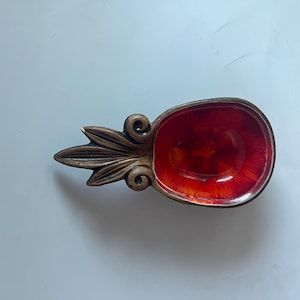 Puede incluir: Un plato decorativo de cerámica con un interior rojo vibrante y un asa marrón en forma de hoja. El plato ovalado es probablemente para servir porciones pequeñas o como elemento decorativo. Hecho a mano.