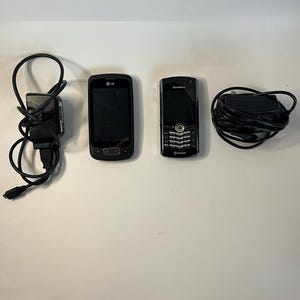 Op de afbeelding: Een assortiment zwarte mobiele telefoons en opladers. De afbeelding toont een LG-telefoon, een Blackberry-telefoon en twee zwarte opladers met opgerolde snoeren. De telefoons worden gepresenteerd op een witte ondergrond.