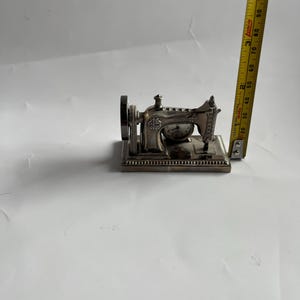 Op de afbeelding: Een miniatuur zilveren naaimachine met ingewikkelde details, waaronder een groot wiel en decoratieve patronen. Een gele rolmaat ligt naast de machine en toont metingen in centimeters.