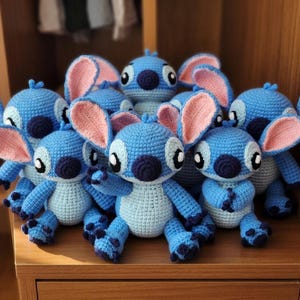 May include: A group of handmade blue crochet dolls with large pink ears, dark blue noses, and black eyes. The dolls are displayed on a wooden surface, highlighting their detailed craftsmanship and whimsical design. The dolls are inspired by the character Stitch.