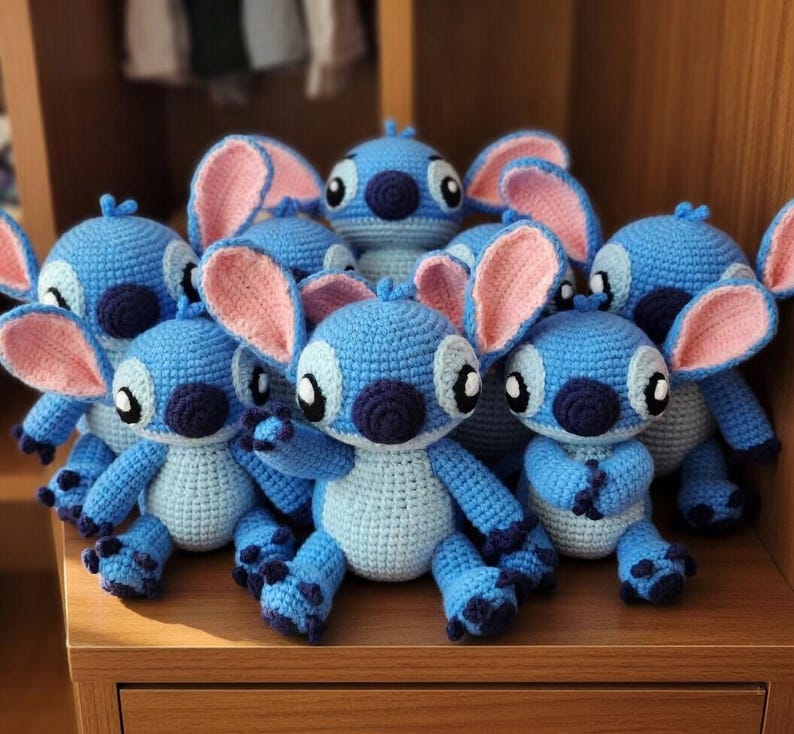 May include: A group of handmade blue crochet dolls with large pink ears, dark blue noses, and black eyes. The dolls are displayed on a wooden surface, highlighting their detailed craftsmanship and whimsical design. The dolls are inspired by the character Stitch.