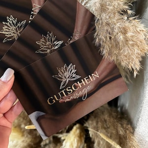 以下が含まれることがあります： ダークブラウンの背景に金箔の蓮の花のアイコンと金色の文字でGUTSCHEIN massageと印刷された長方形のギフトカードの束。カードには、背中に置かれた手の写真画像がデザインされています。