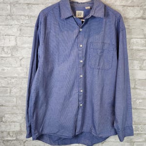 May include: A blue long-sleeve button-down shirt with a chest pocket. The shirt has a collared neckline and white buttons. The brand tag "GAP" is visible on the collar. The shirt is made of a textured fabric.