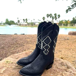 Könnte beinhalten: Ein Paar schwarze Cowboy-Stiefel mit einem strukturierten Muster und silbernen geometrischen Designs am Schaft. Die Stiefel werden im Freien auf einem Felsen präsentiert, mit einem See und Bäumen im Hintergrund.