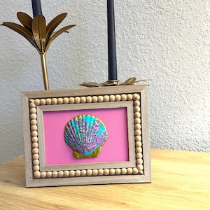 May include: A rectangular wooden picture frame with a beaded border surrounding a bright pink background. Centered on the pink background is a decorative seashell painted in vibrant shades of turquoise, purple, and gold with a textured surface.