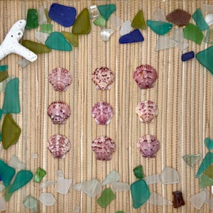 May include: Nine pink and white fan-shaped seashells arranged in a three-by-three grid, surrounded by a border of assorted multi-colored sea glass pieces and two white coral fragments on a light brown textured bamboo mat surface.