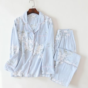 Könnte beinhalten: Hellblaues Pyjama-Set mit floralem Muster. Das Set besteht aus einem Hemd mit Knöpfen, einer Brusttasche und langen Ärmeln sowie einer passenden Hose. Die Kanten des Hemdes und der Manschetten sind rosa eingefasst. Der Pyjama hängt an einem Holzkleiderbügel.