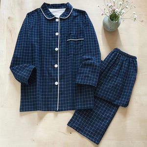 May include: A navy blue plaid pajama set. The long-sleeved button-down shirt has white piping along the collar, cuffs, and pocket. The matching pants have an elastic waistband. The set is made of a soft, comfortable material.