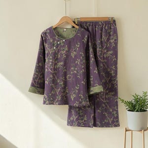 Può includere: Un completo pigiama a due pezzi con motivo floreale. Il set comprende un top a maniche lunghe con scollo tondo e chiusura con bottoni e pantaloni abbinati. Il tessuto è viola scuro con un motivo floreale verde e finiture verdi.