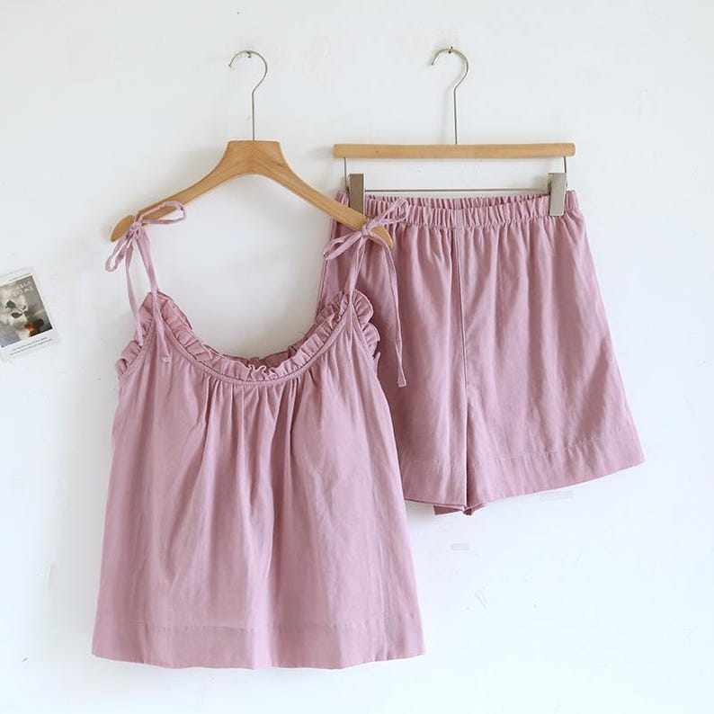 May include: A light pink pajama set, consisting of a camisole top and shorts, hangs on a wooden hanger. The top has tie straps and a ruffled neckline. The shorts have an elastic waistband. The set is made of a soft, lightweight fabric.