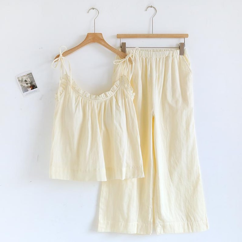May include: A light yellow pajama set, including a camisole top with tie straps and ruffled trim, and matching wide-leg pants. The set is made of a soft fabric and hangs on a wooden hanger against a white background. A small picture is on the wall.