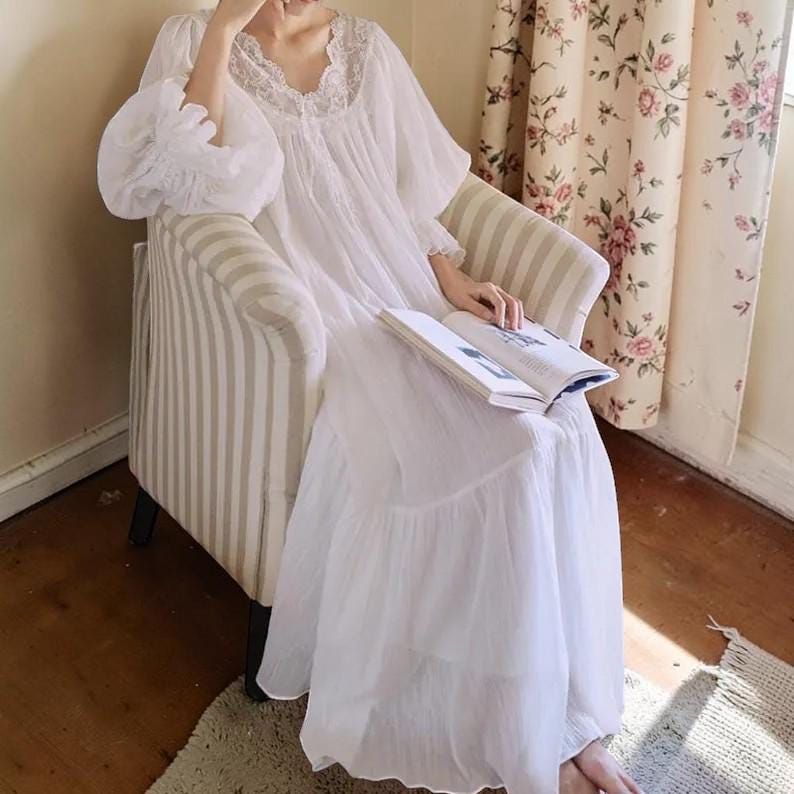 Puede incluir: Un camis&oacute;n blanco de manga larga con ribete de encaje alrededor del escote y las mangas. El camis&oacute;n tiene una falda fluida con volantes y est&aacute; hecho de una tela ligera. La persona est&aacute; sentada en un sill&oacute;n a rayas y sosteniendo un libro.