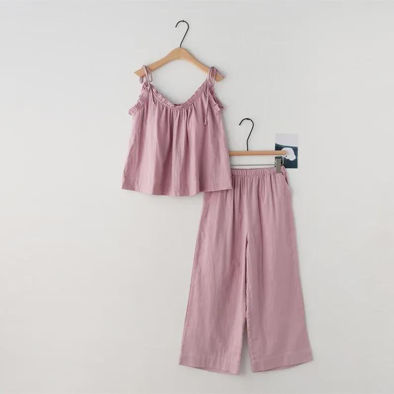 May include: A two-piece pajama set in a soft mauve color. The set includes a camisole top with tie straps and wide-leg pants. Both pieces are hanging on wooden hangers against a plain white background.