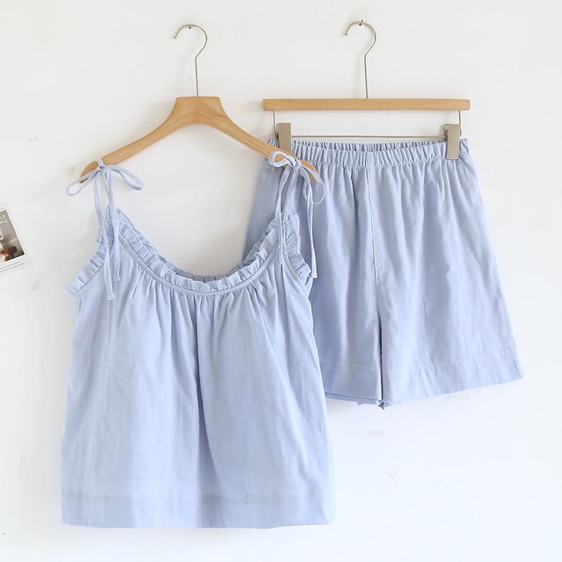 May include: A light blue pajama set, consisting of a camisole top and shorts, hangs on a wooden hanger. The top features tie straps and a ruffled neckline. The shorts have an elastic waistband. The set is made of a soft, lightweight fabric.