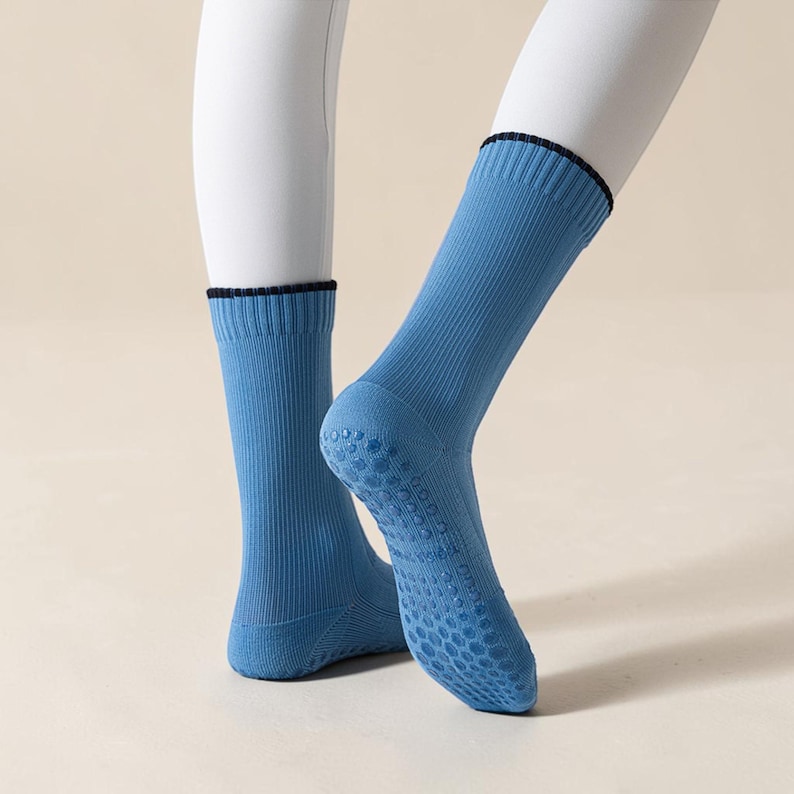 May include: A pair of light blue socks with a ribbed design and a dark blue trim at the top. The soles feature a textured, non-slip pattern. The socks are displayed on a white mannequin leg.