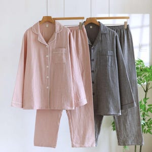 Könnte beinhalten: Zwei Pyjama-Sets hängen an einer Holzstange. Ein Set ist hellrosa mit weißen Verzierungen, das andere grau. Jedes Set enthält ein Hemd mit Knöpfen und einer Brusttasche sowie eine passende Hose. Die Pyjamas sind aus einem weichen, strukturierten Stoff gefertigt.