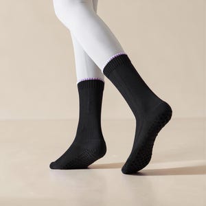 May include: Black crew socks with ribbed texture and a non-slip grip sole. The socks have a white and purple trim at the top. The socks are worn with white leggings.