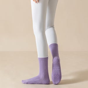 May include: A pair of lavender socks with a white trim at the top. The soles of the socks have a textured, non-slip design. The socks are worn with light gray leggings.