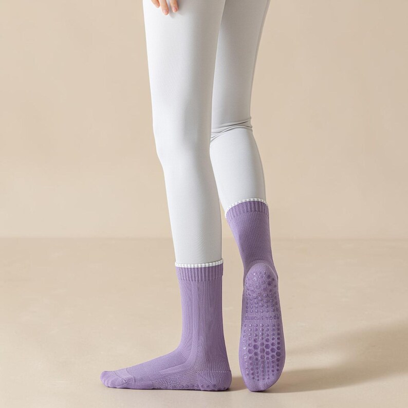 May include: A pair of lavender socks with a white trim at the top. The soles of the socks have a textured, non-slip design. The socks are worn with light gray leggings.