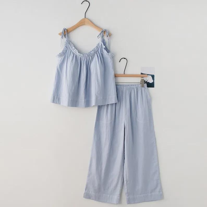 May include: A light blue pajama set, consisting of a camisole top and wide-leg pants, hangs on a wooden hanger. The top features tie straps and a ruffled neckline. The pants have an elastic waistband. The set is made of a soft, lightweight fabric.