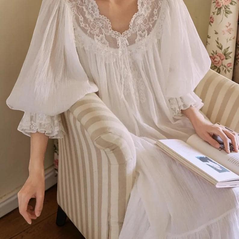 Puede incluir: Un camis&oacute;n blanco de manga larga con detalles de encaje alrededor del escote y los pu&ntilde;os. El camis&oacute;n tiene un cuello en V y una falda fluida y en capas. Las mangas son abullonadas y recogidas en la mu&ntilde;eca. La persona est&aacute; sentada en un sill&oacute;n a rayas y sosteniendo un libro.