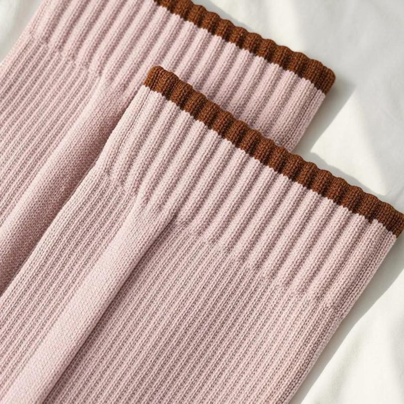 May include: Close-up of two pink ribbed knit socks with a brown scalloped trim. The socks are folded and appear to be made of a soft material. The background is a plain white surface.