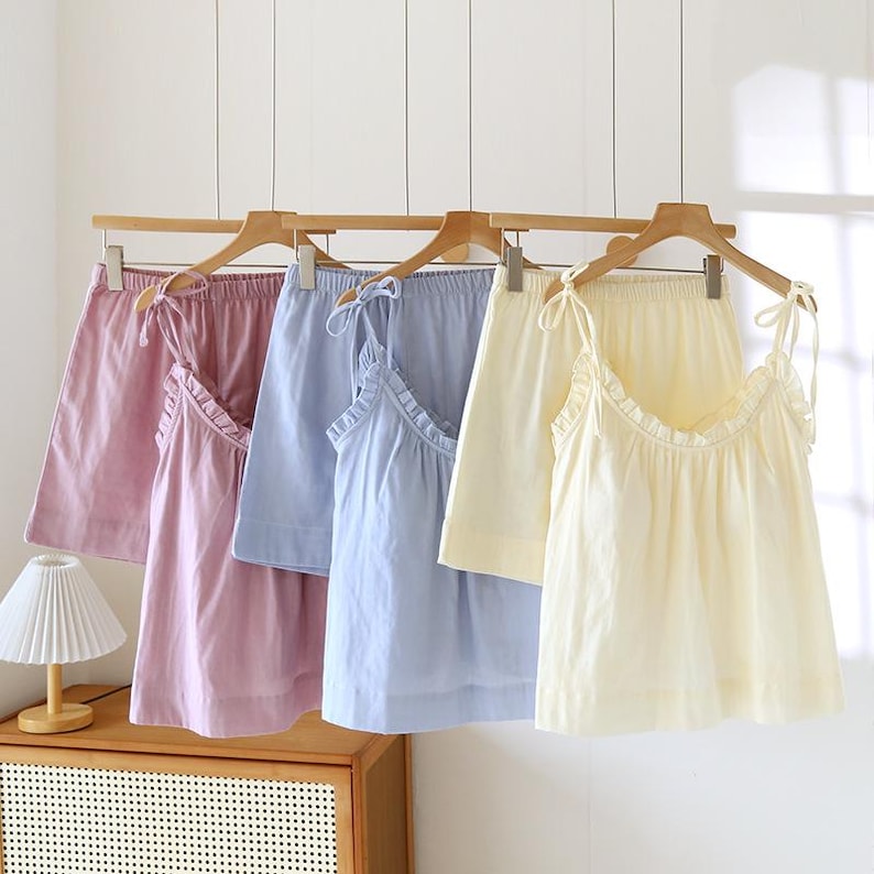 May include: Four sets of pastel-colored pajama sets hanging on wooden hangers. Each set includes a camisole top with ruffled trim and shorts. Colors include pink, light blue, and yellow. The camisoles have tie straps.
