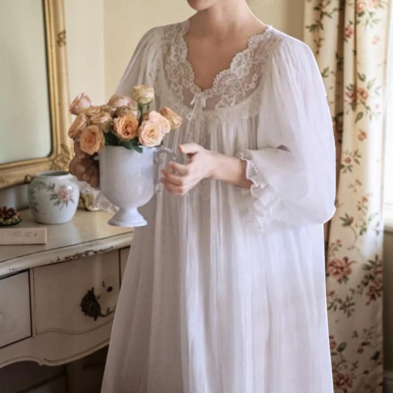 Puede incluir: Un camis&oacute;n blanco con detalles de encaje alrededor del escote y las mangas. La persona sostiene un jarr&oacute;n blanco lleno de rosas color melocot&oacute;n. La escena est&aacute; ambientada en una habitaci&oacute;n con cortinas florales y una c&oacute;moda vintage.