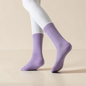May include: A pair of lavender-colored socks with a ribbed design and a white trim at the top. The soles of the socks have a dotted, non-slip grip pattern. The socks are shown on a person wearing white leggings.