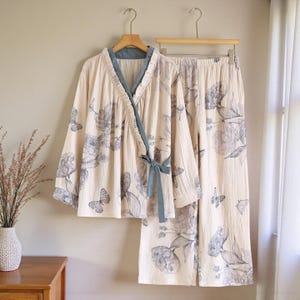 May include: Cream-coloured two-piece pyjama set with a floral and butterfly print. The set includes a wrap-style top with a blue trim and tie, and matching wide-leg trousers. The pyjamas are hanging on a wooden hanger.