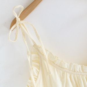 May include: A cream-colored camisole top with tie straps, hanging on a wooden hanger. The top features a ruffled neckline and a soft, flowing fabric. The overall aesthetic is simple and elegant, suitable for warm weather.
