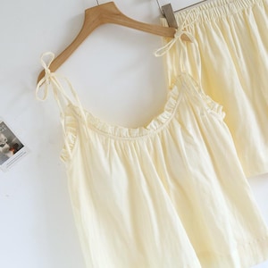 May include: A light yellow pajama set, including a camisole top and shorts, hangs on a wooden hanger. The camisole features delicate tie straps and ruffled edges. The shorts have an elastic waistband. The set is made of a soft, lightweight fabric.