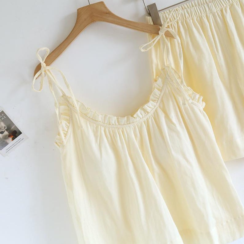 May include: A light yellow pajama set, including a camisole top and shorts, hangs on a wooden hanger. The camisole features delicate tie straps and ruffled edges. The shorts have an elastic waistband. The set is made of a soft, lightweight fabric.