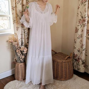 Puede incluir: Camis&oacute;n largo blanco con escote en V y detalles de encaje. El camis&oacute;n presenta mangas con volantes y una falda escalonada. La imagen muestra el camis&oacute;n en una habitaci&oacute;n con cortinas florales y una cesta de mimbre.