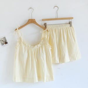 May include: A two-piece pajama set in a soft, pale yellow color. The set includes a camisole top with ruffled trim and tie straps, and matching shorts with an elastic waistband. Both pieces are hanging on wooden hangers against a white background.