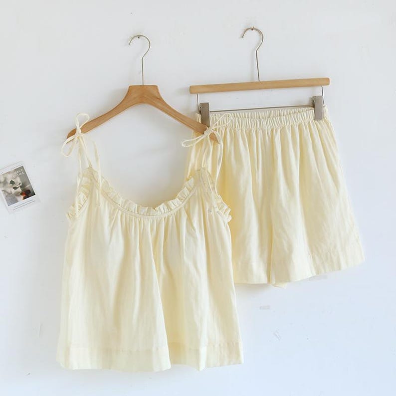 May include: A two-piece pajama set in a soft, pale yellow color. The set includes a camisole top with ruffled trim and tie straps, and matching shorts with an elastic waistband. Both pieces are hanging on wooden hangers against a white background.