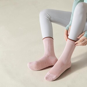 May include: A pair of pink socks with a brown trim at the top and a textured sole. The socks are ribbed and appear to be made of a soft material. The socks are being worn by a person wearing gray leggings.