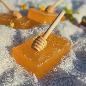Könnte beinhalten: Nahaufnahme von mehreren rechteckigen, bernsteinfarbenen Seifenstücken mit hölzernen Honigwaben. Die Seifenstücke liegen auf einer weißen, flauschigen Oberfläche, mit ein paar gelben Blumen im Hintergrund. Die Honigwaben liegen auf den Seifen.