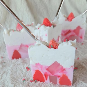 Puede incluir: Tres jabones decorativos que se asemejan a un pastel de fresas. Cada jabón tiene una parte superior blanca, una capa intermedia rosa y un diseño de fresa roja en la parte inferior. Pequeñas chispas en forma de fresa están esparcidas alrededor de los jabones.