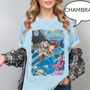 Könnte beinhalten: Hellblaues Chambray-T-Shirt mit einem farbenfrohen Aufdruck von Buzz Lightyear, Woody und anderen Charakteren aus dem Film Toy Story. Der Aufdruck enthält den Text "Buzz Lightyear Laser Blast".