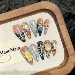 Puede incluir: Un conjunto de diez uñas artificiales con diversos diseños. Las uñas presentan una combinación de colores, incluyendo rosa, azul y dorado, con adornos como cruces, corazones y figuras religiosas. Las uñas están dispuestas sobre una bandeja de madera.