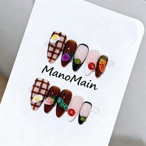 Puede incluir: Un conjunto de uñas artificiales en forma de almendra con varios diseños temáticos de comida. Las uñas presentan decoraciones en miniatura de huevo, verdura y fruta, incluyendo un huevo frito, una zanahoria y una fresa. El texto "ManoMain" está impreso en el fondo blanco.
