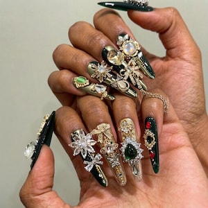 Puede incluir: Uñas postizas largas y puntiagudas de color verde bosque oscuro, decoradas con un surtido de dijes de metal dorado, cristales transparentes, perlas de imitación y pequeños cabujones verdes. Los adornos incluyen lazos y flores.