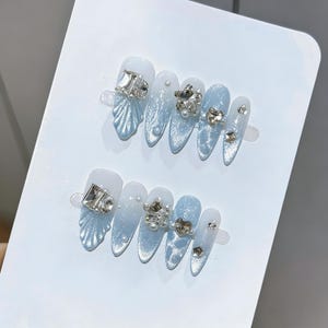 Puede incluir: Dos juegos de uñas postizas con un diseño degradado azul claro y blanco. Cada uña está adornada con pedrería plateada, perlas y elementos decorativos. Las uñas son de forma ovalada y se presentan sobre una tarjeta blanca.