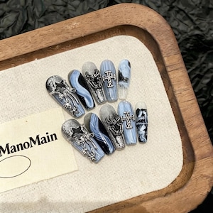 Puede incluir: Un conjunto de uñas postizas con forma de ataúd y temática gótica. Las uñas presentan purpurina plateada, remolinos azules y adornos decorativos, incluyendo cruces, estrellas y diseños de telarañas. La marca "ManoMain" es visible en una etiqueta blanca.